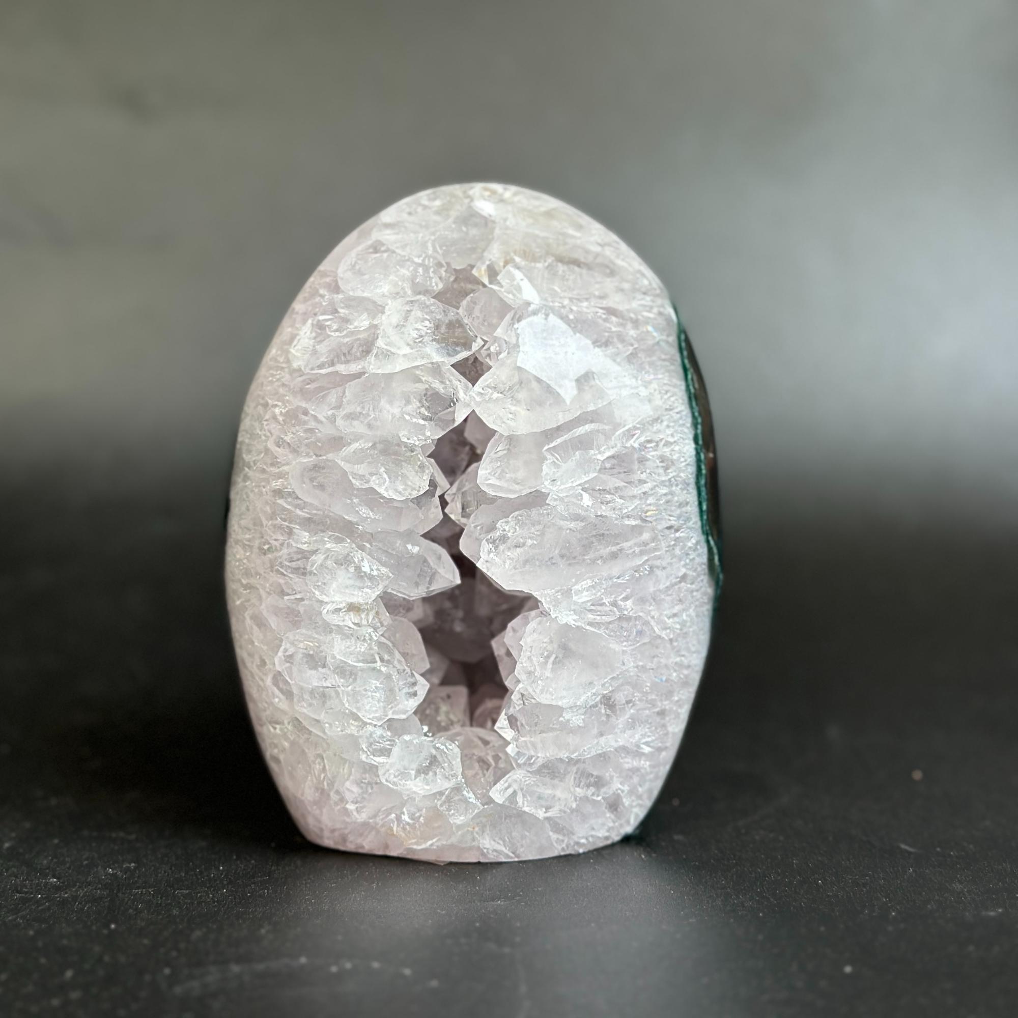 A polished geode-shaped crystal stone with a white and pale pink outer cavity and a dark interior, photographed on a dark gray surface against a gray background.