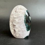 A polished oval mineral specimen with a pale translucent crystal-like exterior and a dark green to black geode cavity visible on one side, photographed on a dark gray surface against a soft gray background.