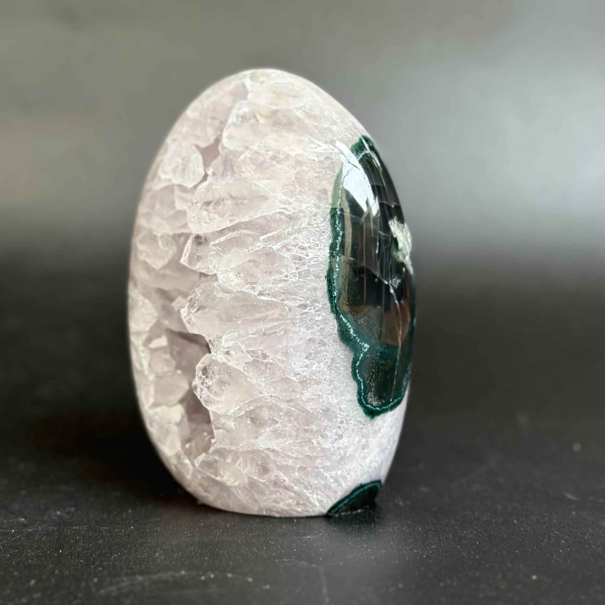 A polished oval mineral specimen with a pale translucent crystal-like exterior and a dark green to black geode cavity visible on one side, photographed on a dark gray surface against a soft gray background.