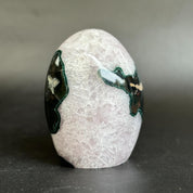 A polished oval stone sculpture with a pale pinkish-white textured surface and dark green and black inlay details on both sides, photographed on a dark gray surface against a gray background.