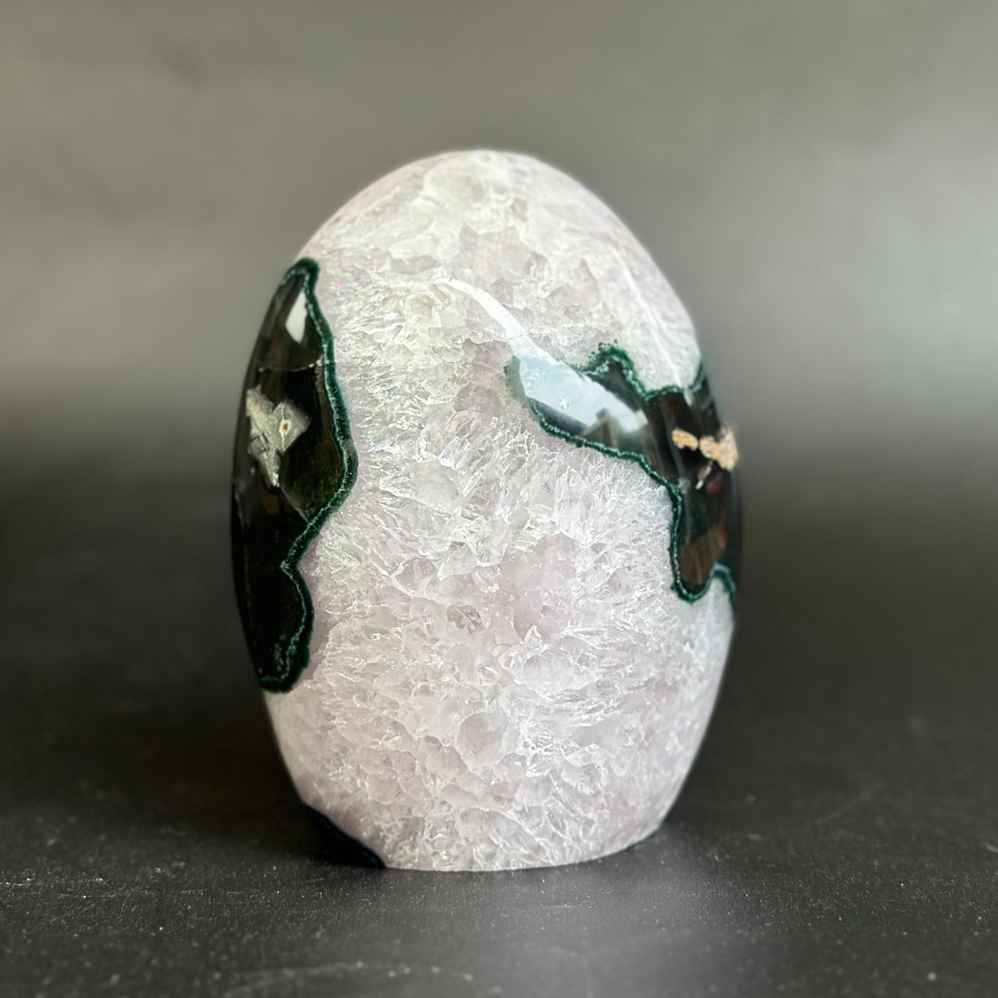 A polished oval stone sculpture with a pale pinkish-white textured surface and dark green and black inlay details on both sides, photographed on a dark gray surface against a gray background.