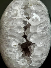 A polished oval stone or crystal cluster with a rough, jagged white and clear quartz geode-like texture, photographed close-up against a dark background.