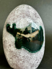 A polished oval stone featuring a light lavender-gray crystalline body with a prominent dark green agate inlay and a thin gold edge, photographed on a dark gray background.