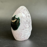 A polished rose quartz crystal tower with a natural black and green agate geode inlay, standing upright on a dark gray surface against a neutral gray background.
