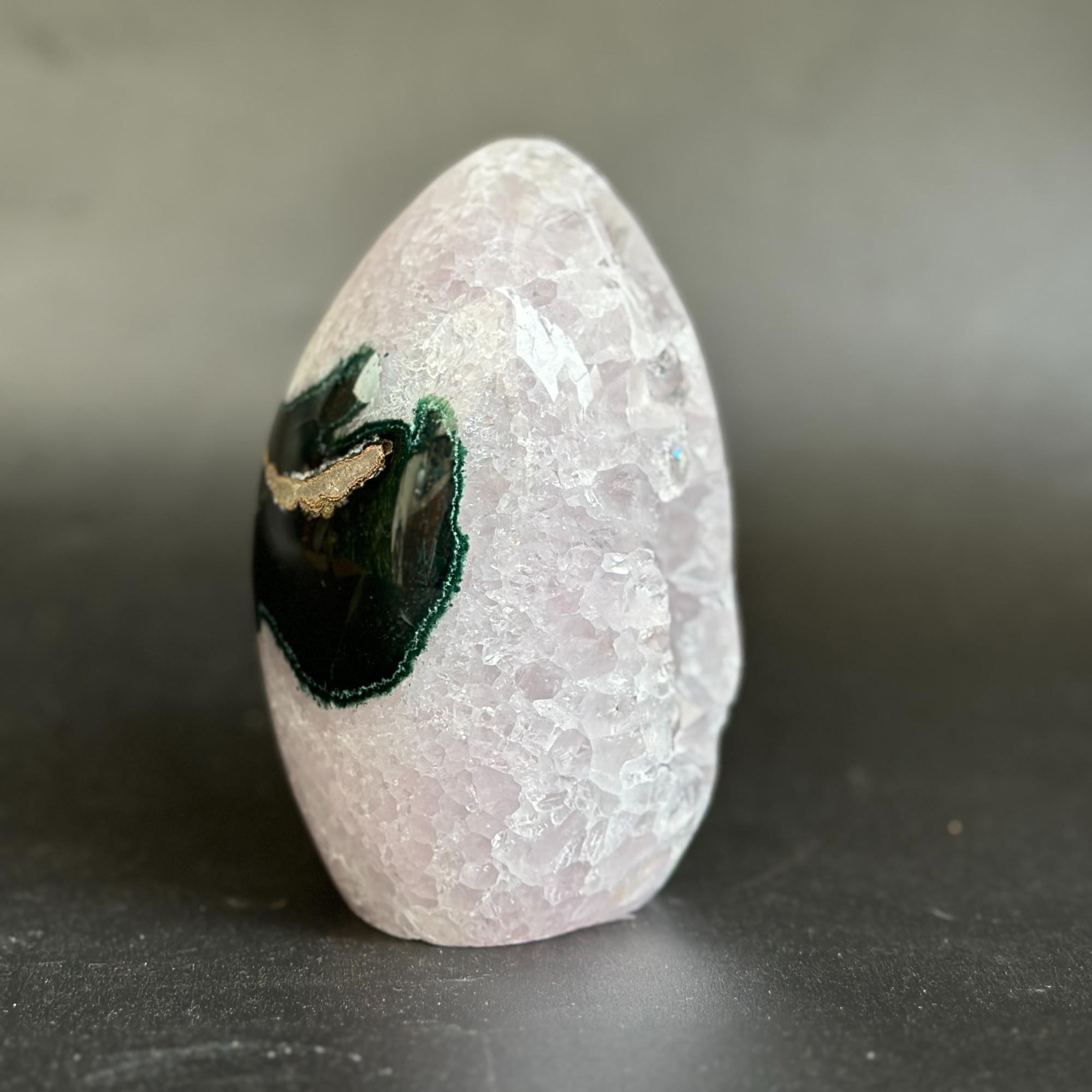 A polished rose quartz crystal tower with a natural black and green agate geode inlay, standing upright on a dark gray surface against a neutral gray background.
