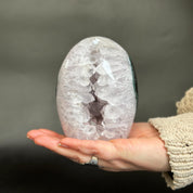A person's hand holding a polished geode specimen with a white and pale lavender crystal interior and a dark outer shell, set against a dark gray background.