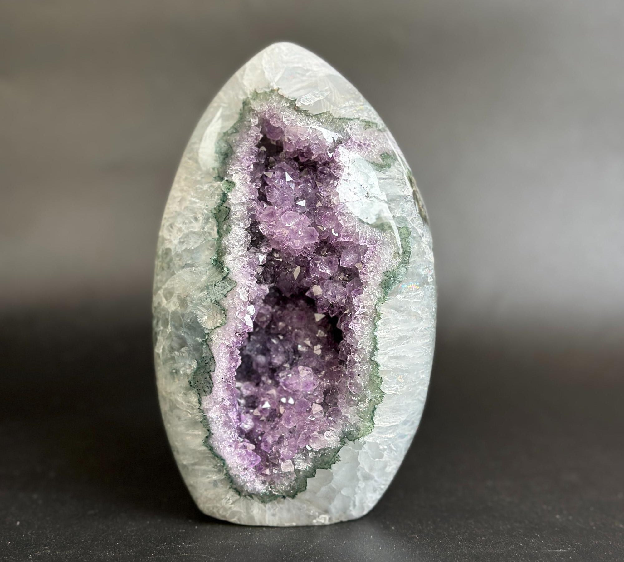 A polished geode sculpture featuring a purple amethyst cavity surrounded by a green and gray crystalline outer layer, set against a dark gray background.