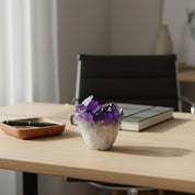 Amethyst crystal in a clear container on a wooden desk with a book and pen in the background.