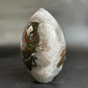A polished teardrop-shaped crystal stone with natural inclusions, featuring a translucent white and gray body with dark brown, black, and subtle purple inclusions, standing on a dark surface.