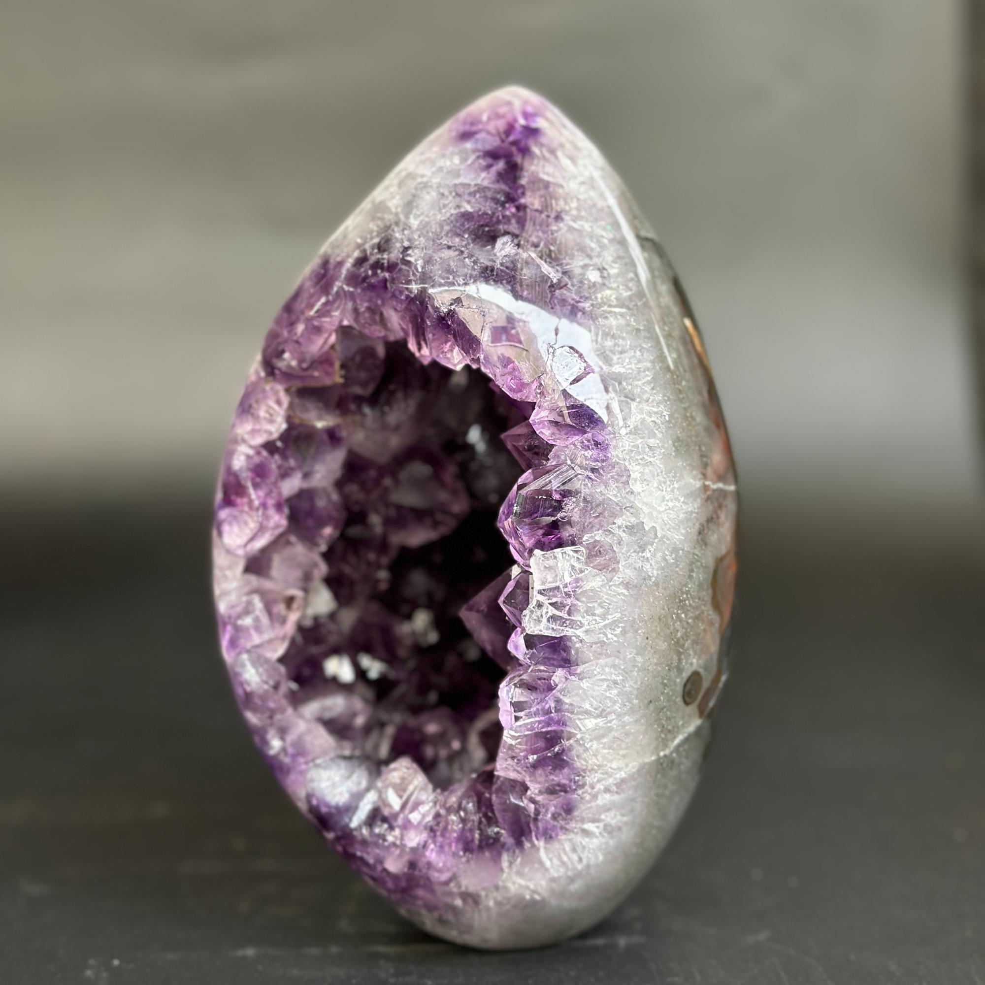A polished teardrop-shaped amethyst geode sculpture with a smooth silver-gray outer surface and a deep purple crystal interior, standing upright on a dark surface against a blurred gray background.