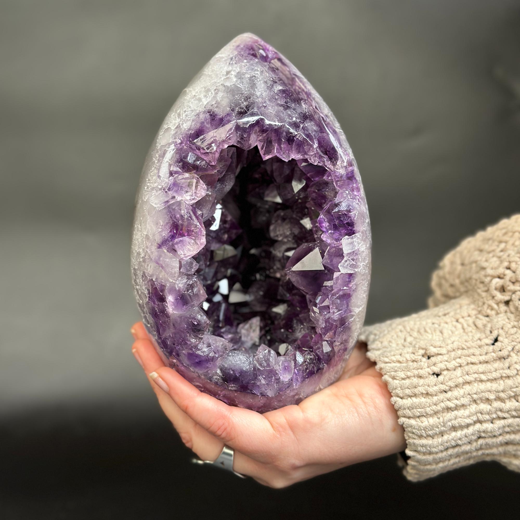 A hand holding a polished teardrop-shaped amethyst geode with a deep purple crystal interior against a dark gray background.