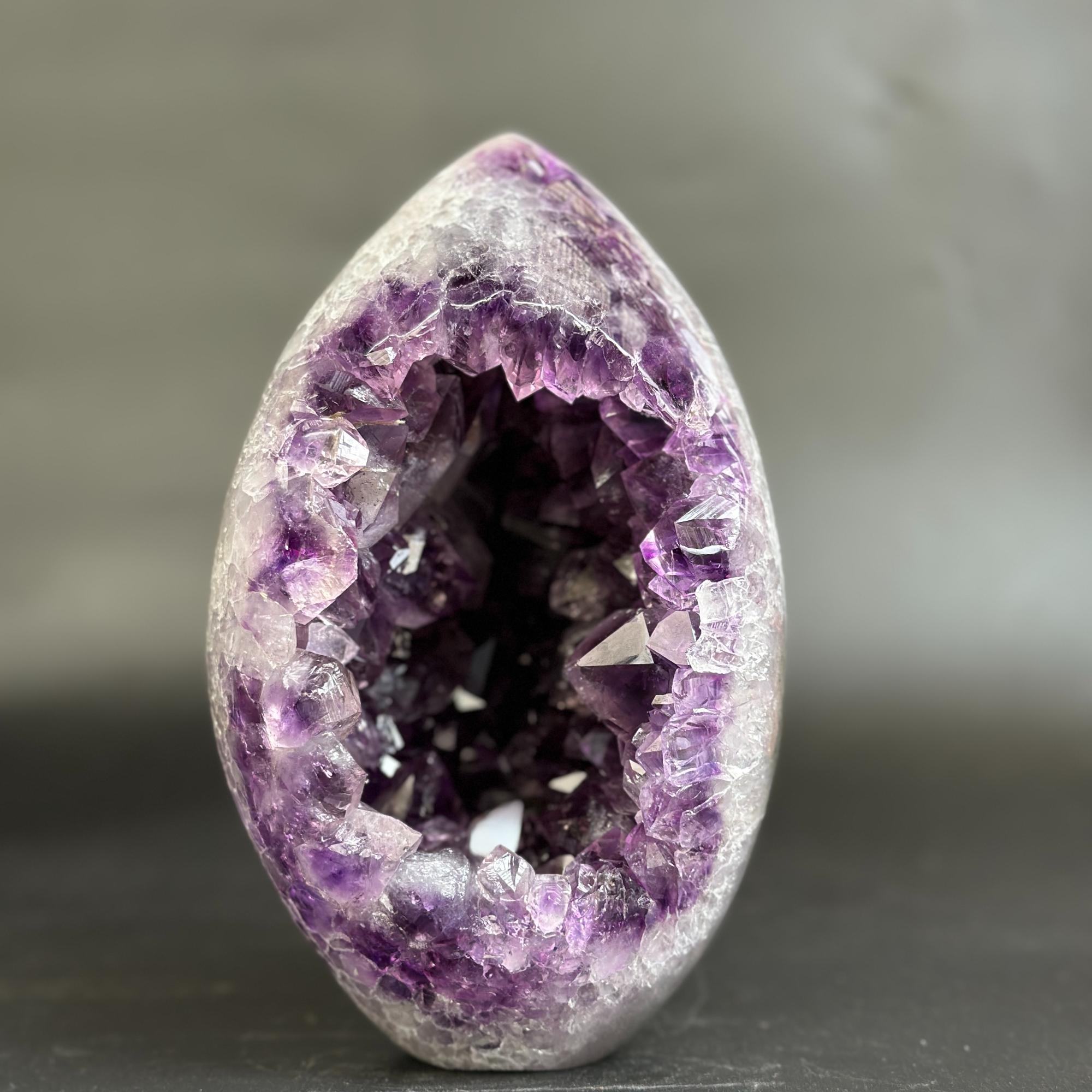 A polished amethyst geode specimen with a smooth teardrop-shaped outer layer and a deep, crystal-filled interior, photographed against a soft gray blurred background.
