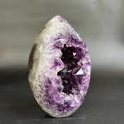 A egg-shaped amethyst geode crystal sculpture with a polished white and purple outer surface and a deep purple crystal cavity, photographed on a dark gray surface against a blurred gray background.