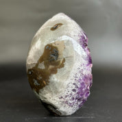 A polished natural crystal stone with a rounded teardrop shape, featuring a translucent milky white body with prominent purple amethyst clusters on one end and dark brown inclusions on the other, set against a dark gray background.