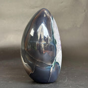 A polished teardrop-shaped agate crystal sculpture with a glossy, highly reflective surface, showcasing dark blue, black, and white natural banding, set against a neutral gray background.