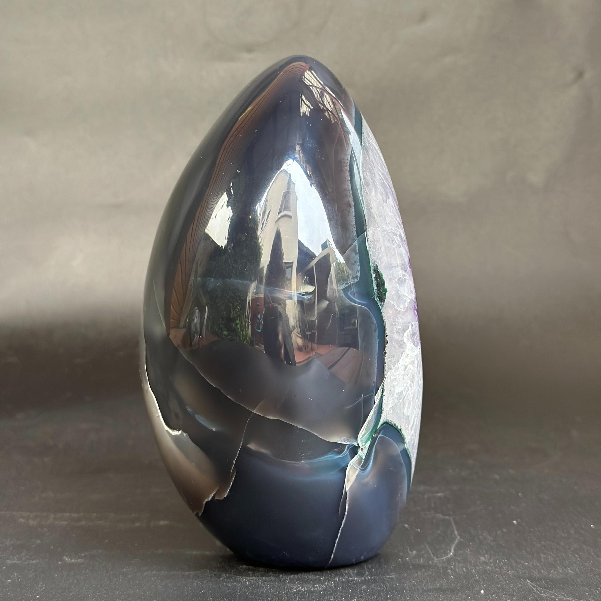 A polished teardrop-shaped agate crystal sculpture with a glossy, highly reflective surface, showcasing dark blue, black, and white natural banding, set against a neutral gray background.