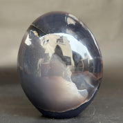 A polished dark agate crystal egg sculpture with a highly reflective, mirror-like surface, showing a reflection of the room and a natural fracture line, set against a neutral gray background.