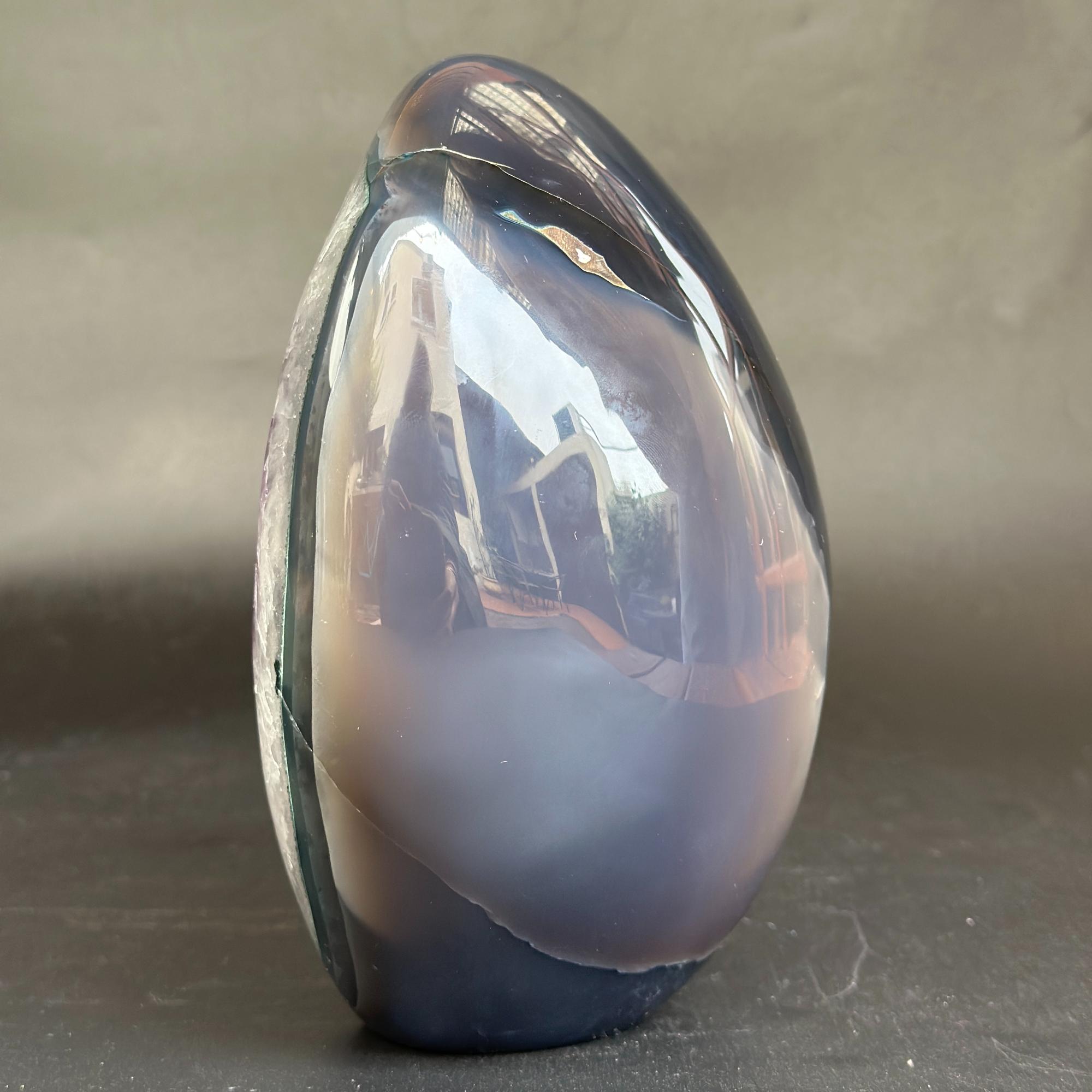 A polished oval agate crystal stone with a smooth, reflective surface, showcasing bands of dark blue, gray, and soft lavender-purple, standing upright on a dark surface against a neutral gray background.