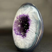 A polished oval agate stone featuring a natural purple amethyst geode cavity surrounded by a white crystalline outer ring and a dark gray outer edge, photographed on a dark gray surface with a blurred gray background.