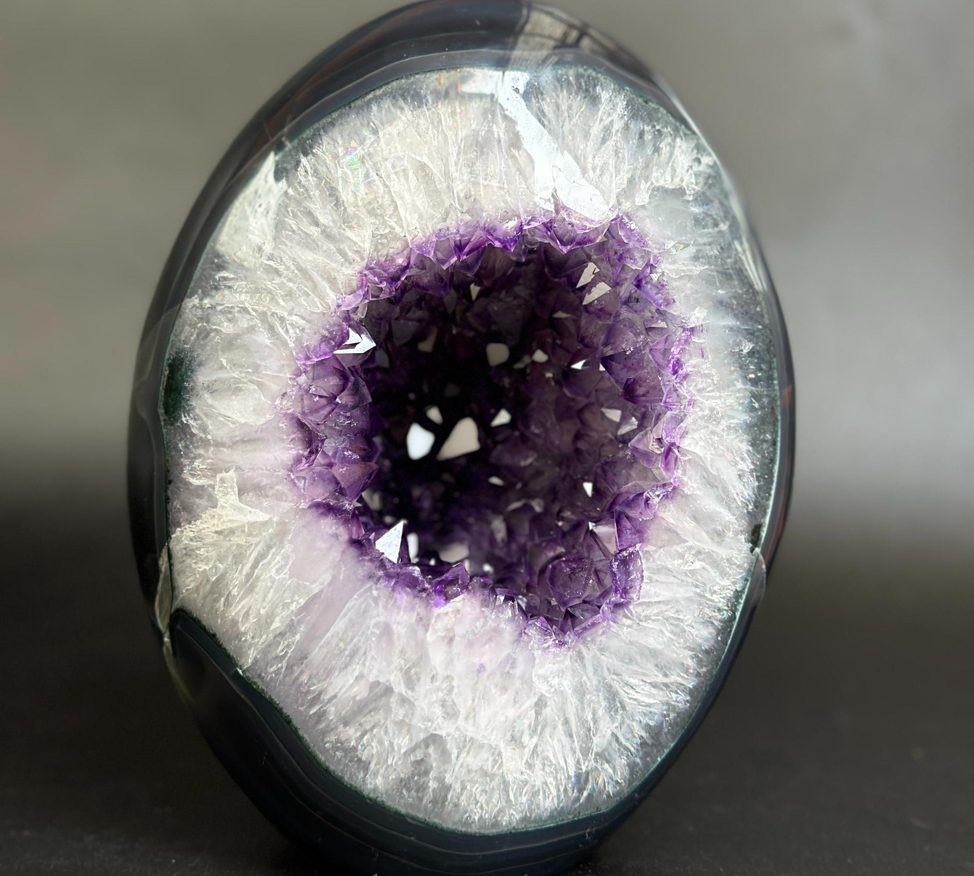 A polished geode-style decorative object with a dark outer shell and a bright white crystalline interior surrounding a deep purple crystal cavity, photographed on a dark neutral background.