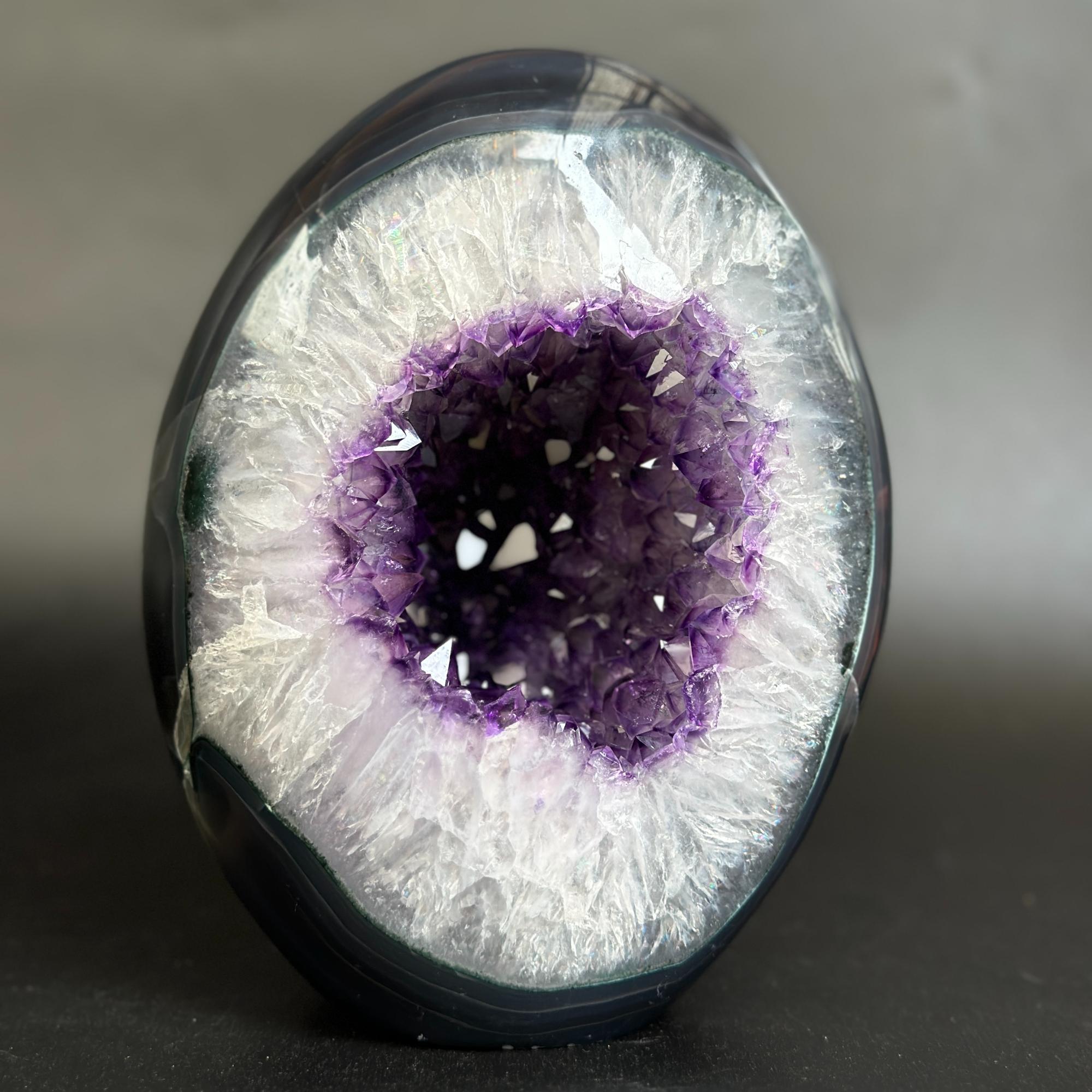 A polished geode-style decorative object with a dark outer shell and a bright white crystalline interior surrounding a deep purple crystal cavity, photographed on a dark neutral background.