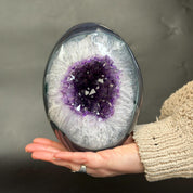A polished geode crystal sphere with a white and purple amethyst interior, held in a person's hand against a dark gray background.