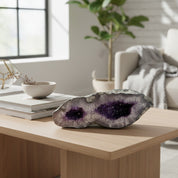 A natural amethyst geode with dark purple crystal cavities and a gray outer matrix, displayed on a light wood coffee table in a modern, sunlit living room.
