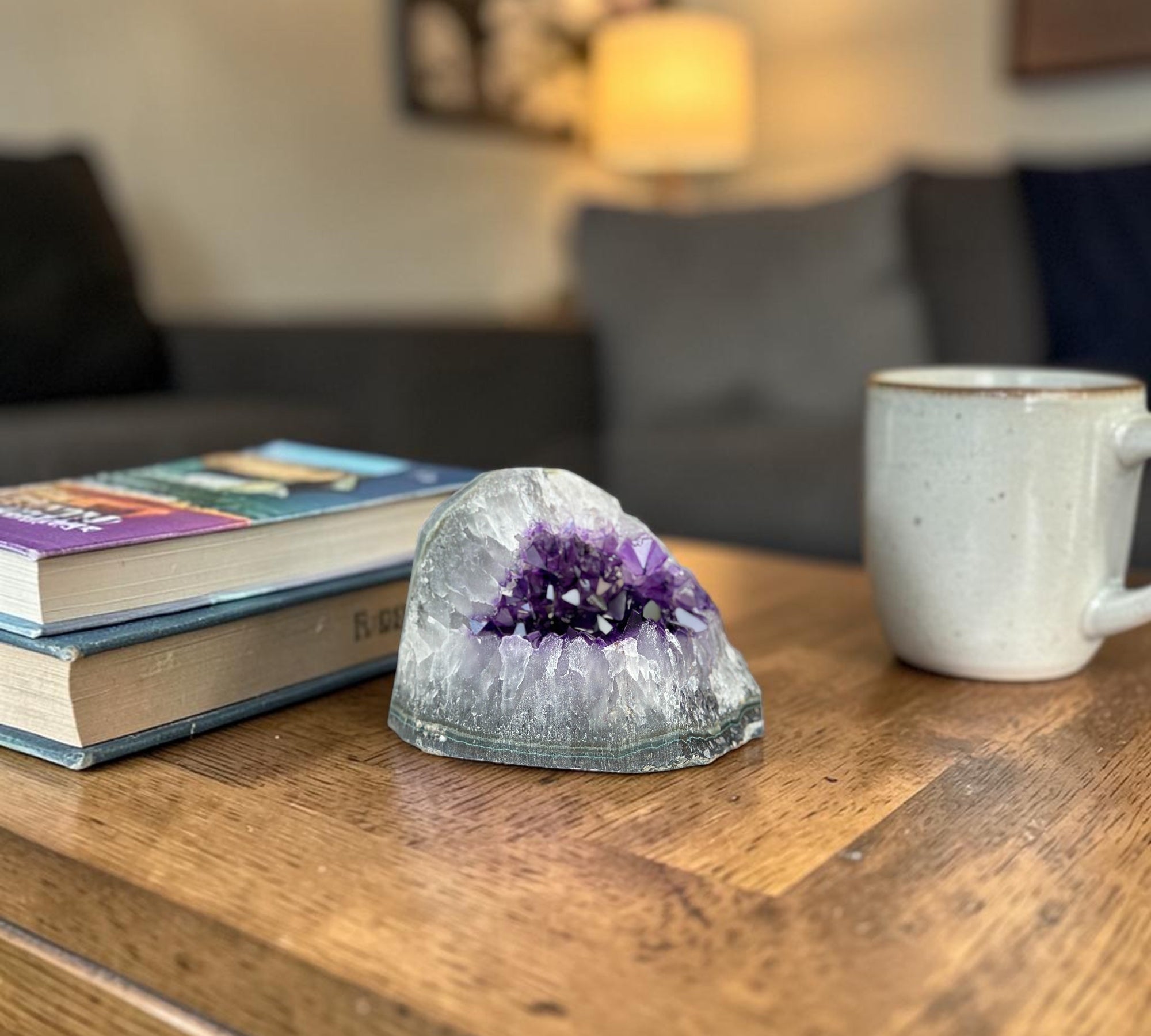 Large, deep purple amethyst geode cluster with a gray exterior, displayed on a wooden table next to books and a mug.