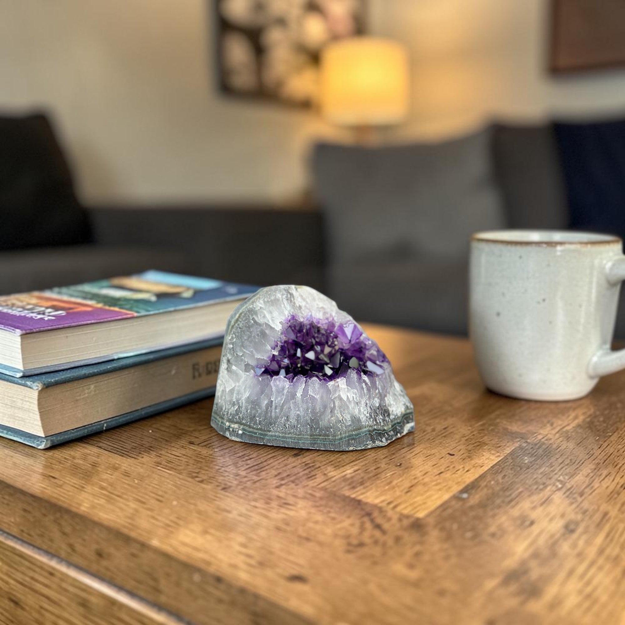 Large, deep purple amethyst geode cluster with a gray exterior, displayed on a wooden table next to books and a mug.