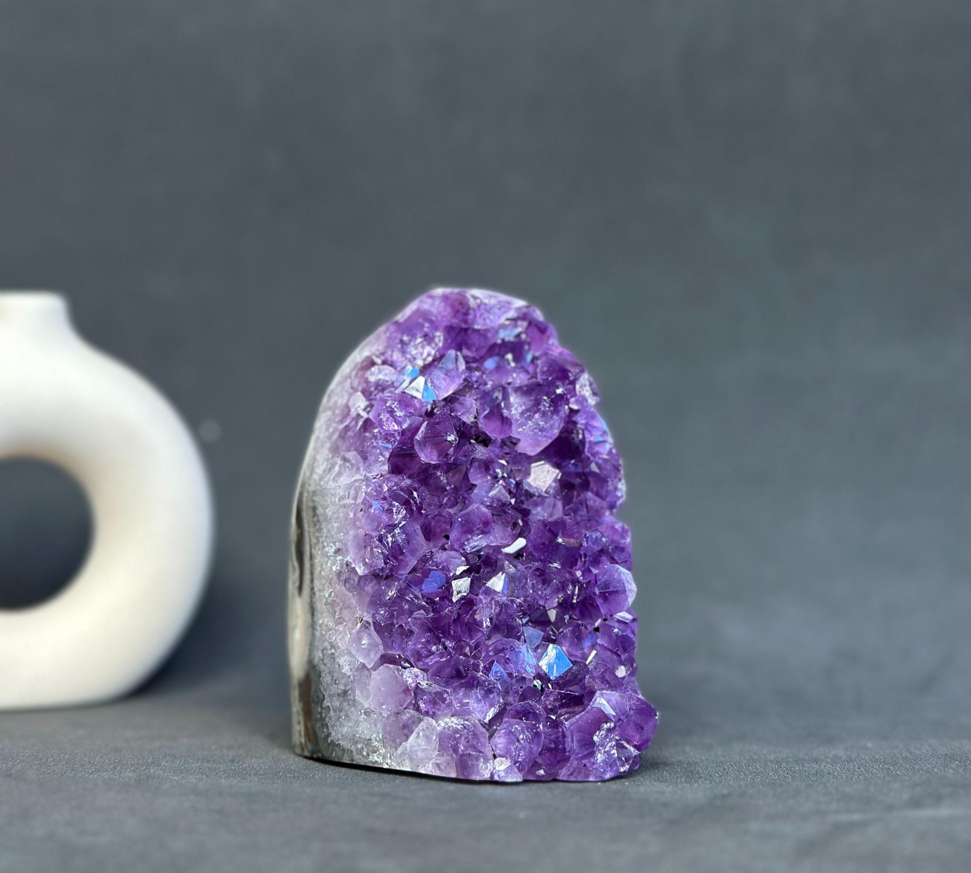 A raw amethyst crystal cluster geode with a natural rough stone base, displayed on a dark gray surface with a blurred white ceramic object in the background.