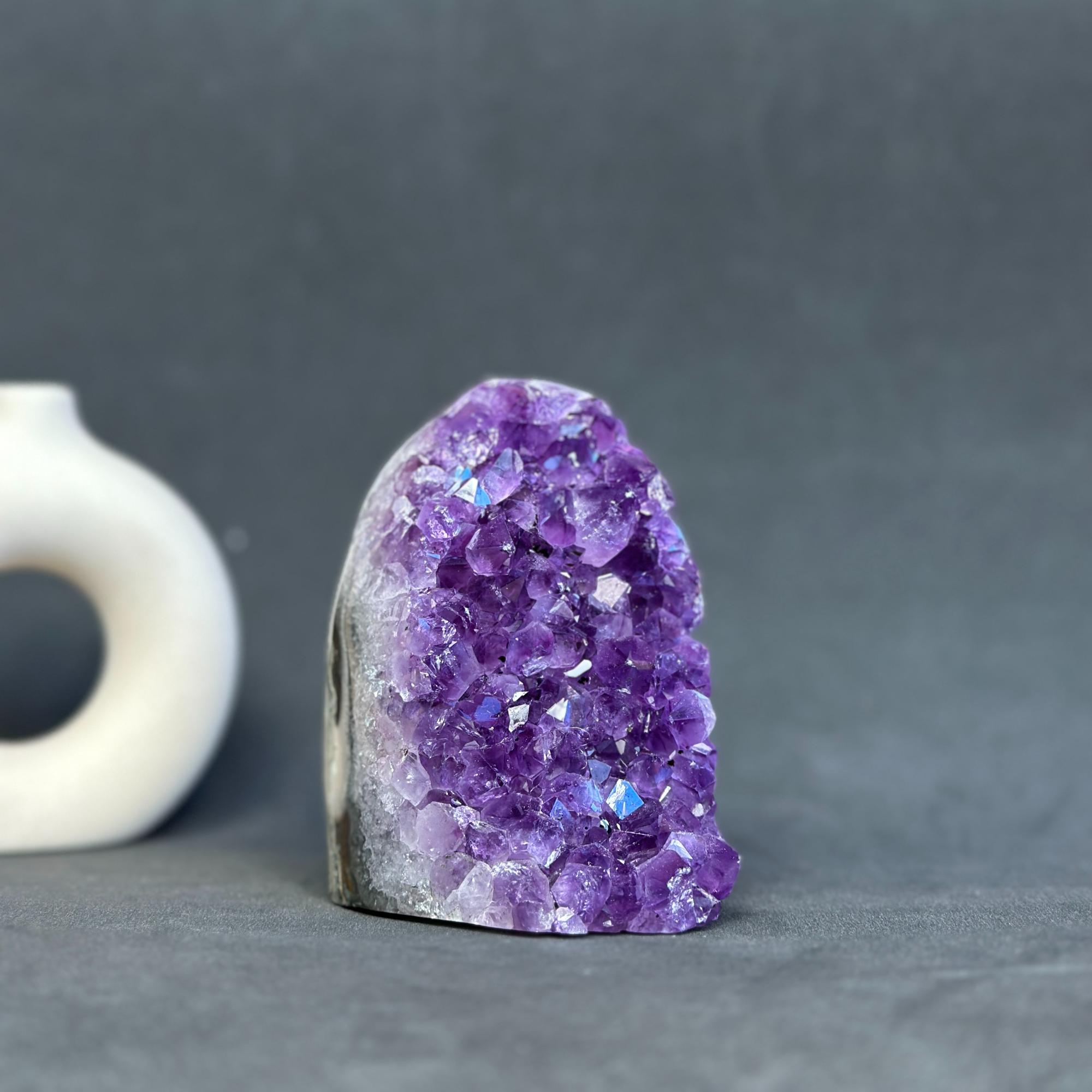 A raw amethyst crystal cluster geode with a natural rough stone base, displayed on a dark gray surface with a blurred white ceramic object in the background.