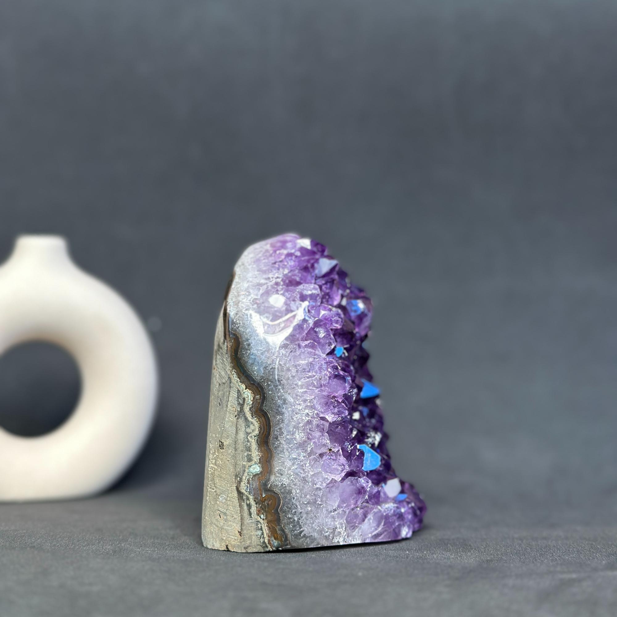 A natural amethyst geode specimen with purple crystal points and a gray-brown matrix base, photographed on a dark gray surface with a small white object blurred in the background.