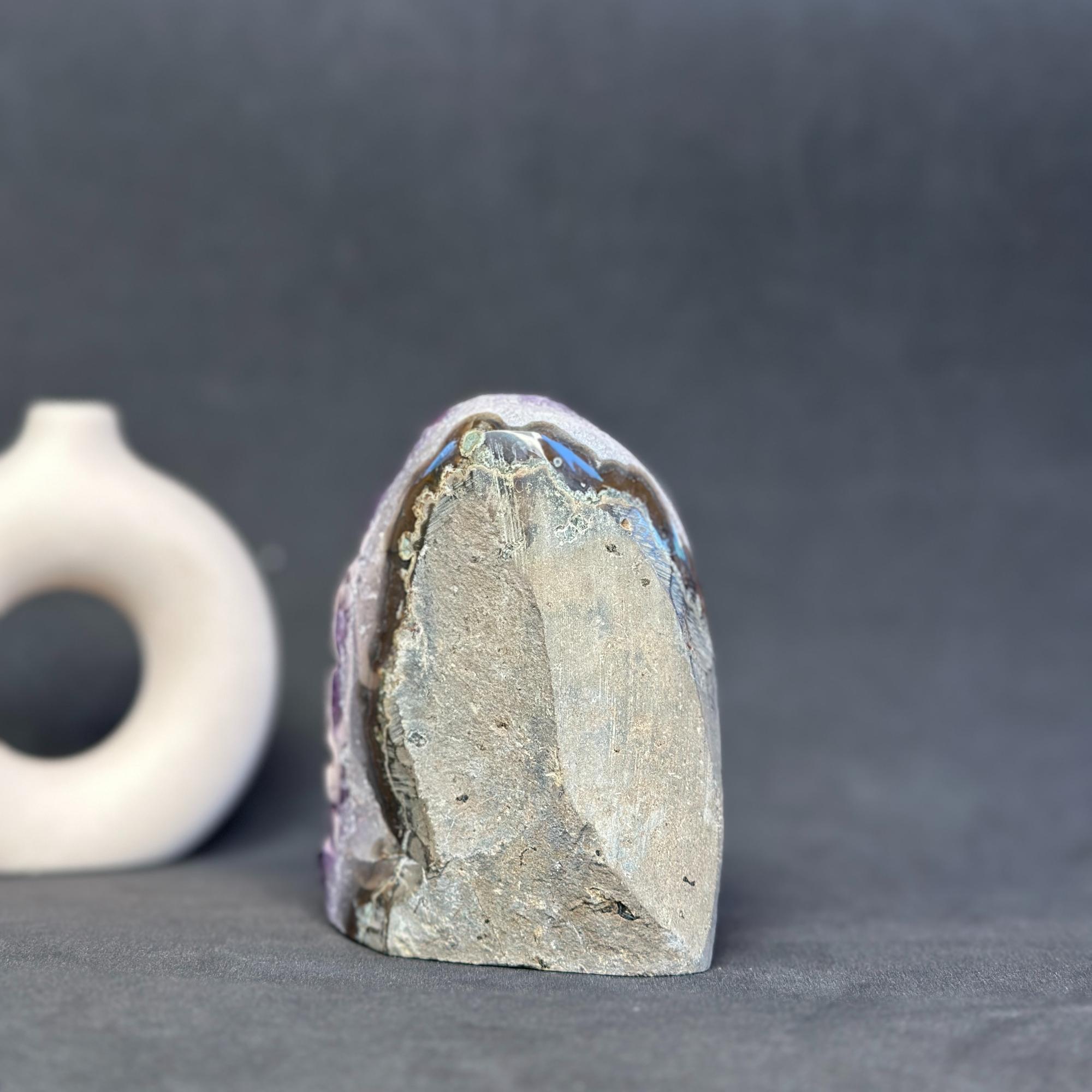 A raw, rough-hewn crystal or mineral specimen with a gray and beige textured surface and subtle iridescent blue-green highlights, standing upright on a dark gray surface. A small white ring-shaped object is partially visible in the blurred background on the left.