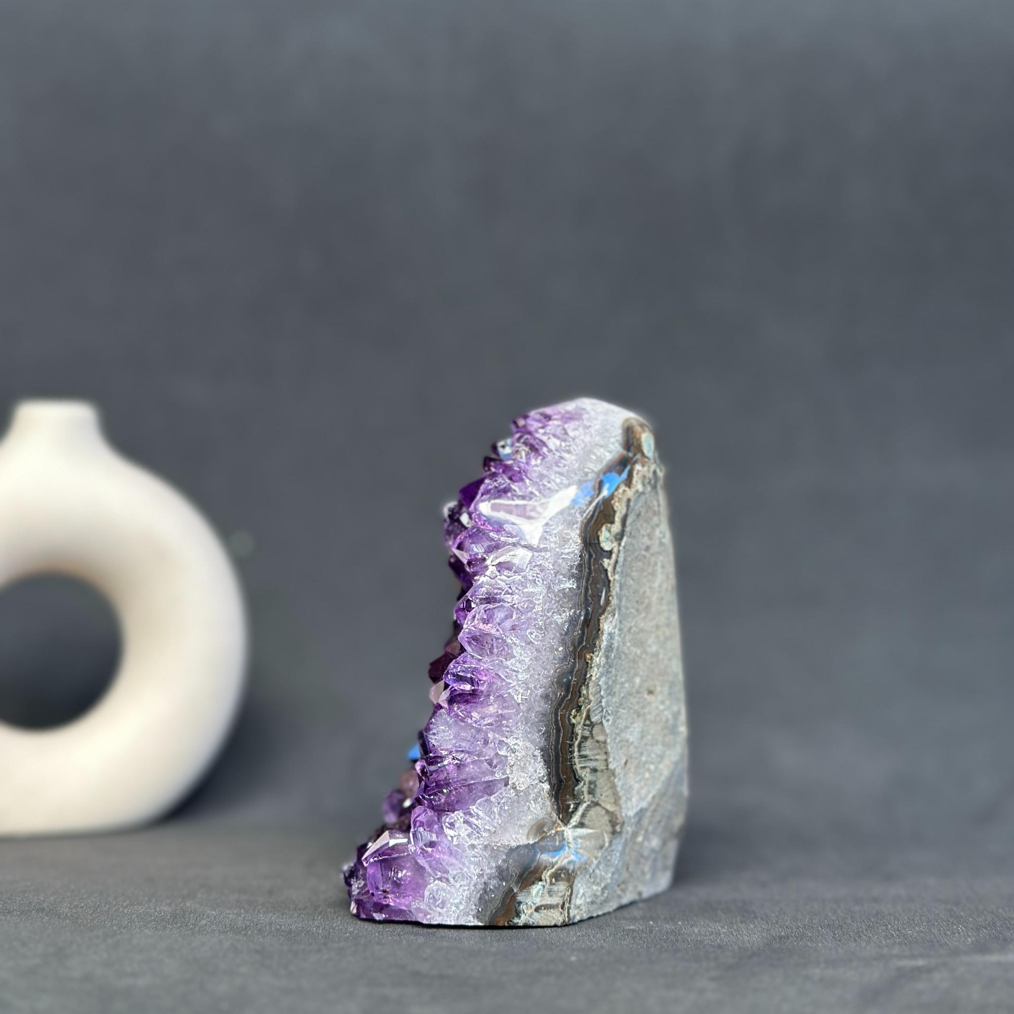 A raw amethyst geode specimen with purple crystal points and a gray matrix base, displayed on a dark gray surface with a small white ceramic object blurred in the background.