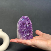 A hand holding a purple amethyst crystal cluster geode against a dark gray background.