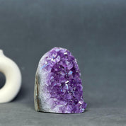 A natural amethyst crystal cluster with a rough, polished dark gray and brown base, photographed on a dark gray surface with a blurred white object in the background.