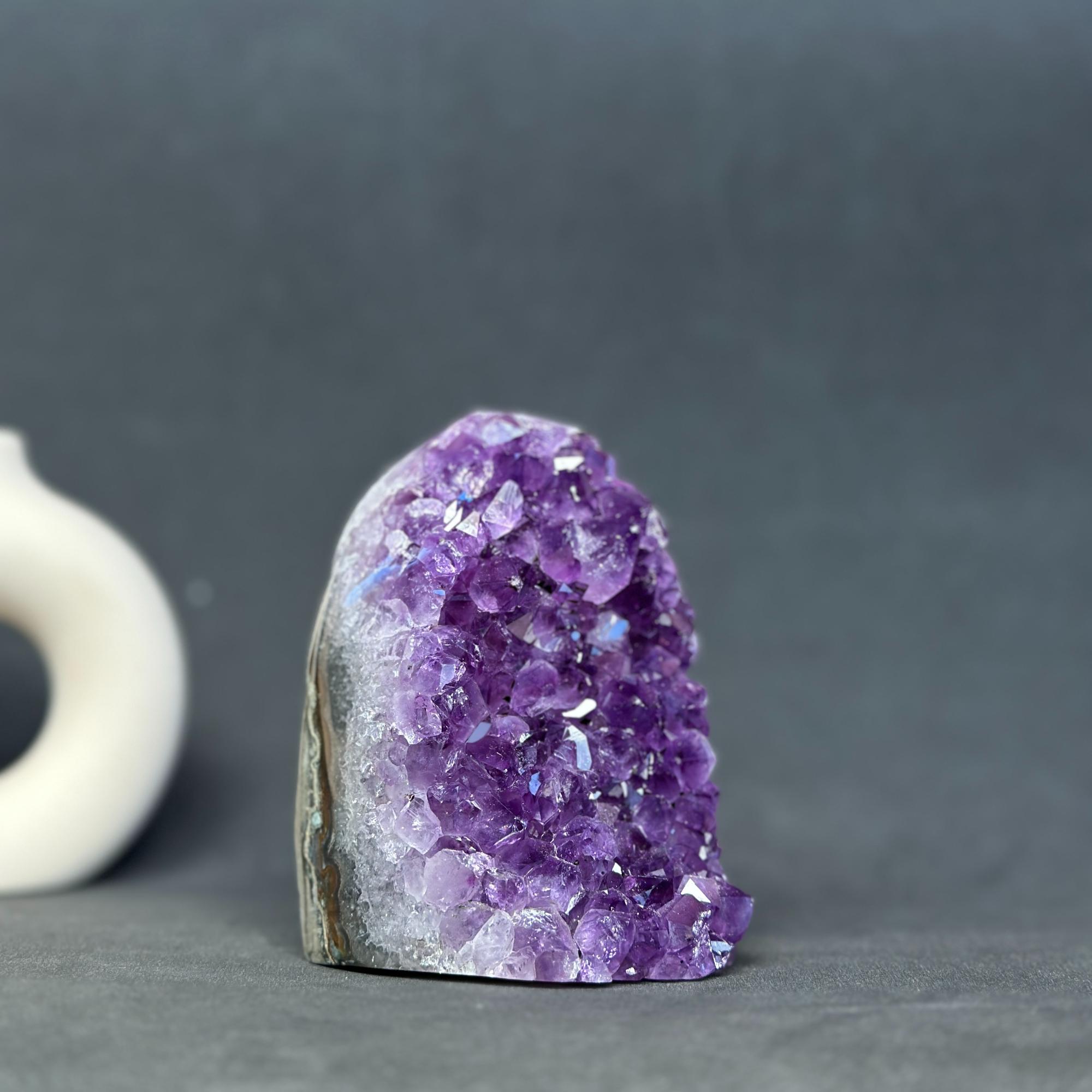 A natural amethyst crystal cluster with a rough, polished dark gray and brown base, photographed on a dark gray surface with a blurred white object in the background.