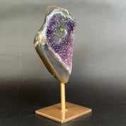 A polished agate geode mount with a purple crystal cavity, displayed on a gold-colored metal stand against a dark gray background.