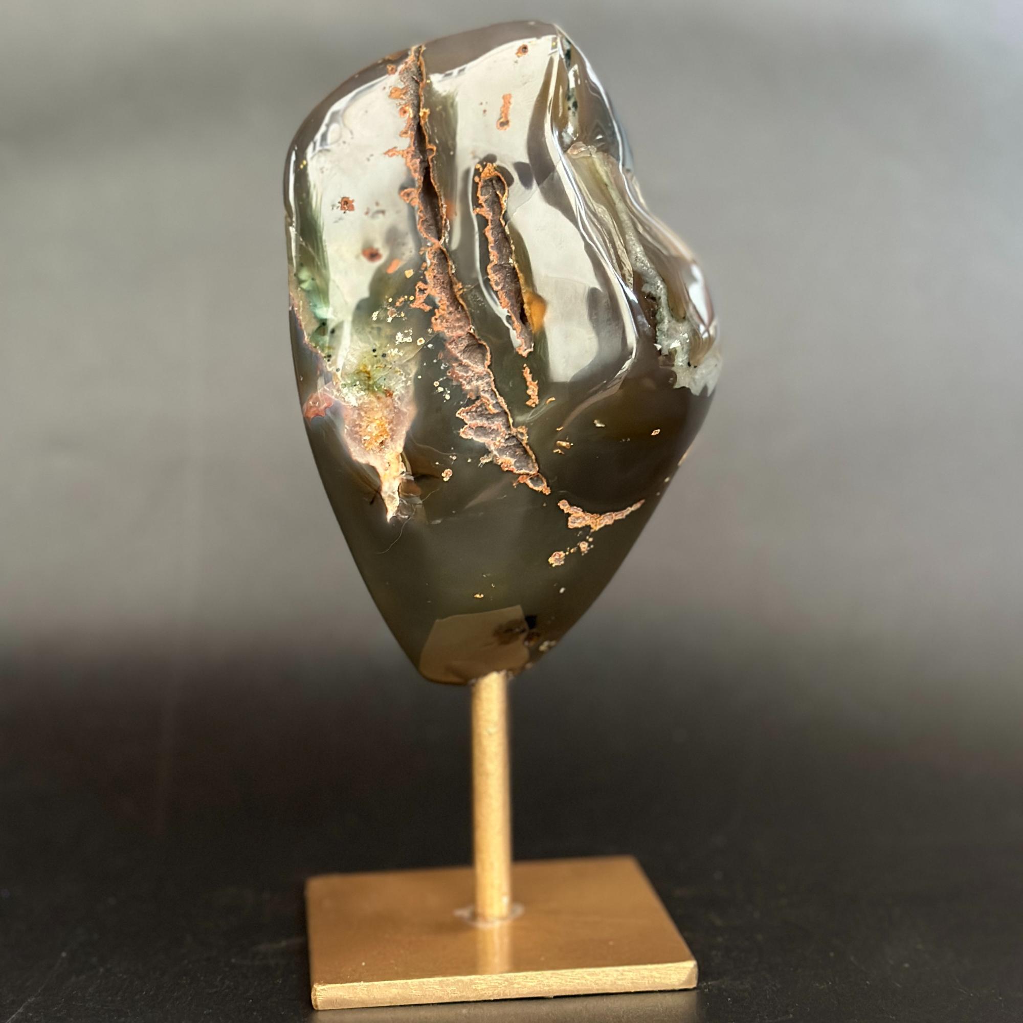A polished dark agate or jasper stone with copper and green inclusions, mounted on a small wooden display stand against a dark gray background.