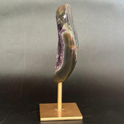 A polished agate geode slice with a natural purple crystal cavity, mounted on a light wood display stand against a dark gray background.