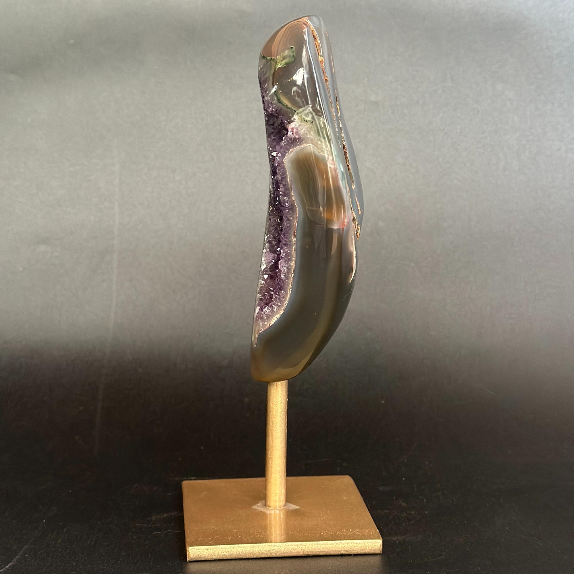 A polished agate geode slice with a natural purple crystal cavity, mounted on a light wood display stand against a dark gray background.