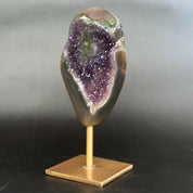 A polished natural crystal geode mount with a purple amethyst interior and smooth gray-brown outer layers, displayed on a small gold-colored metal stand against a dark gray gradient background.