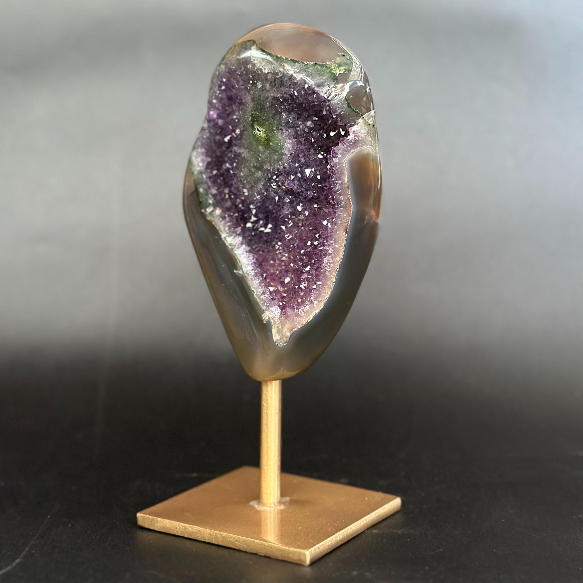 A polished natural crystal geode mount with a purple amethyst interior and smooth gray-brown outer layers, displayed on a small gold-colored metal stand against a dark gray gradient background.