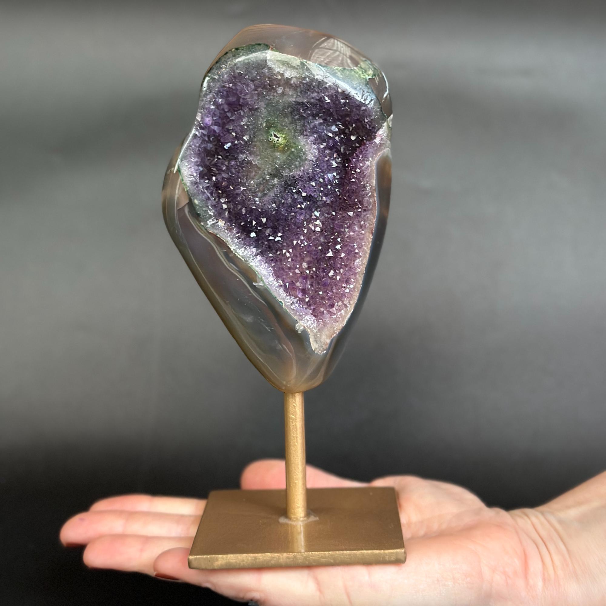 A polished natural amethyst geode display mounted on a brass stand, held in the open palm of a hand against a dark gray background.
