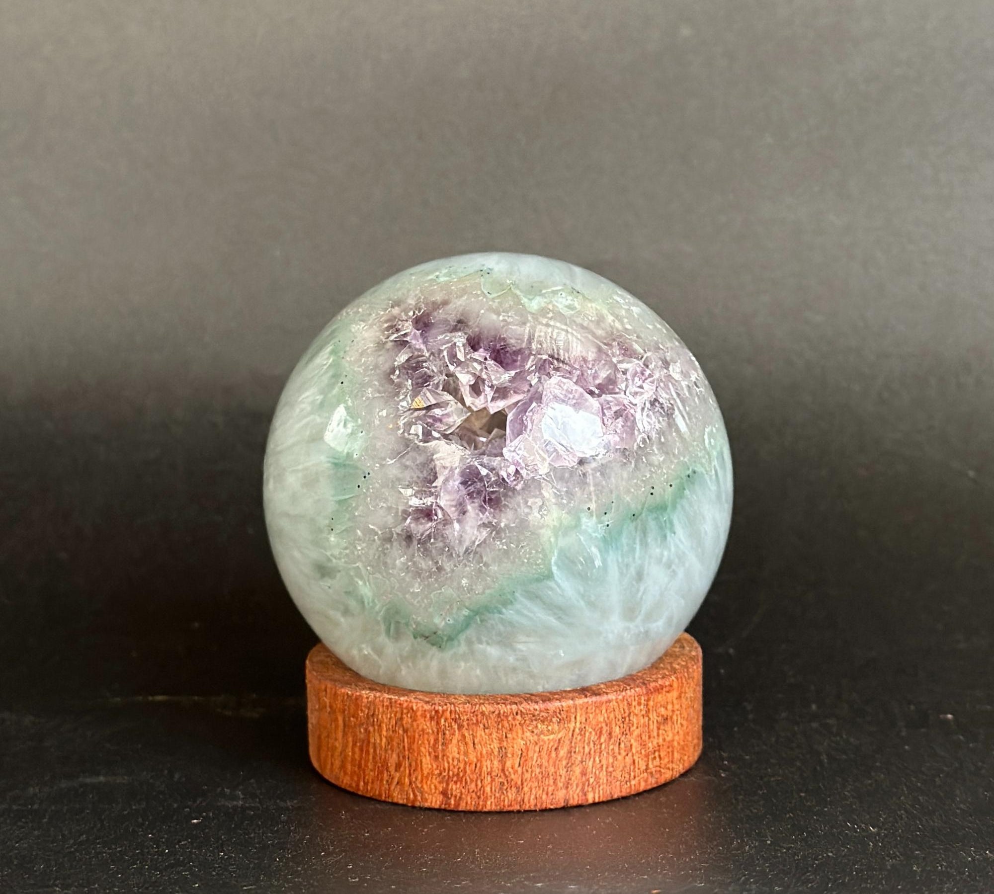A polished crystal geode sphere with purple and green mineral clusters, displayed on a round wooden stand against a dark gray background.