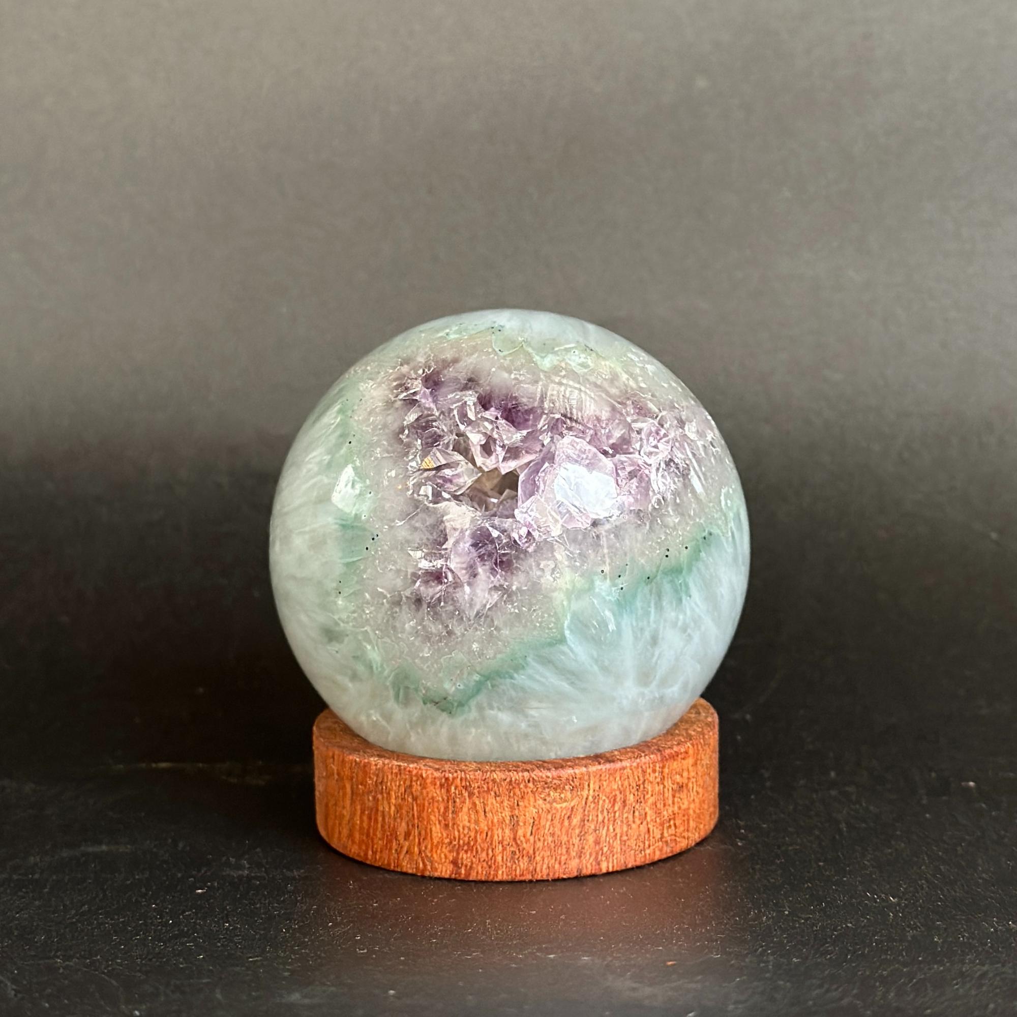 A polished crystal geode sphere with purple and green mineral clusters, displayed on a round wooden stand against a dark gray background.