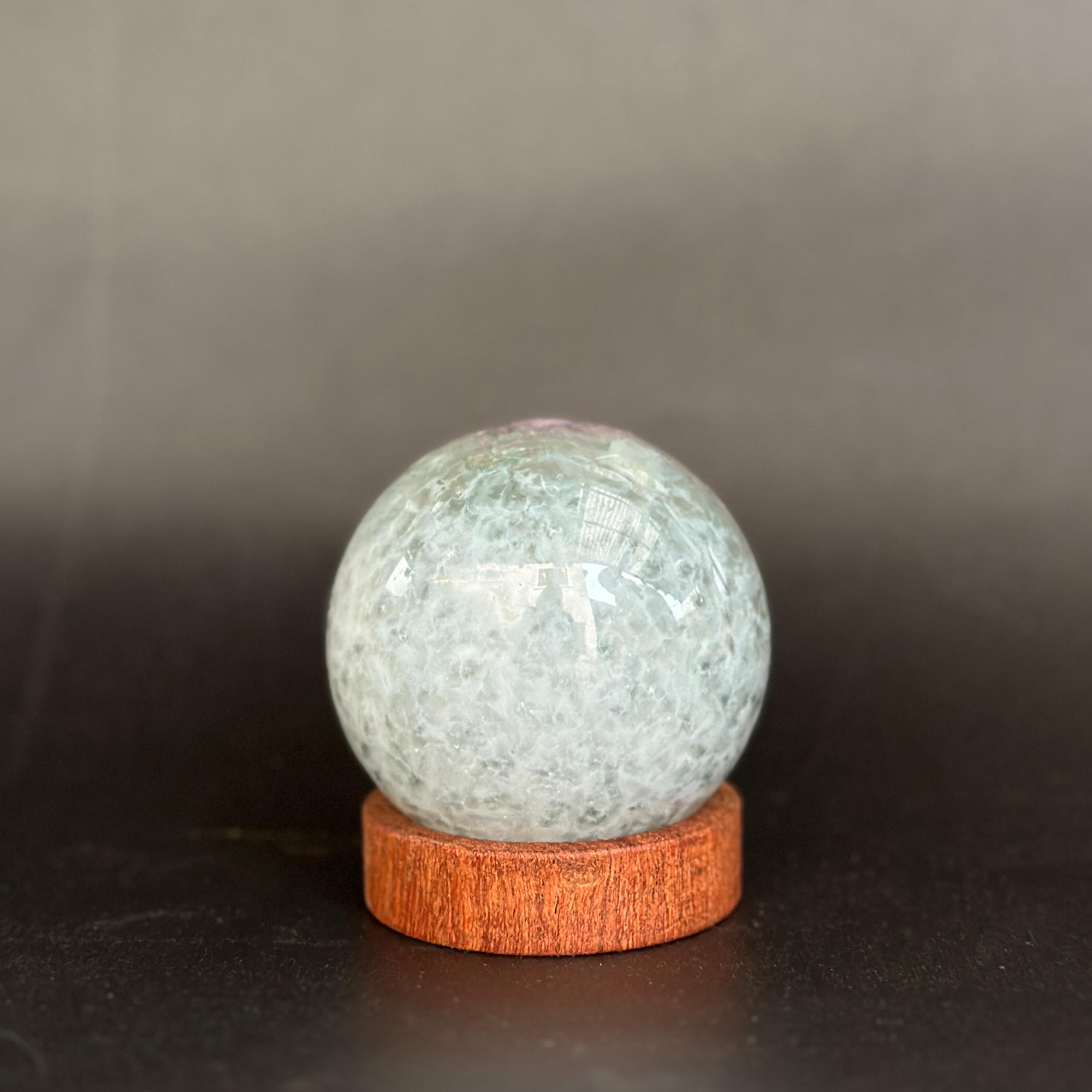 A round light blue-green crystal ball with white cloudy inclusions, displayed on a small round wooden base against a dark gray background.