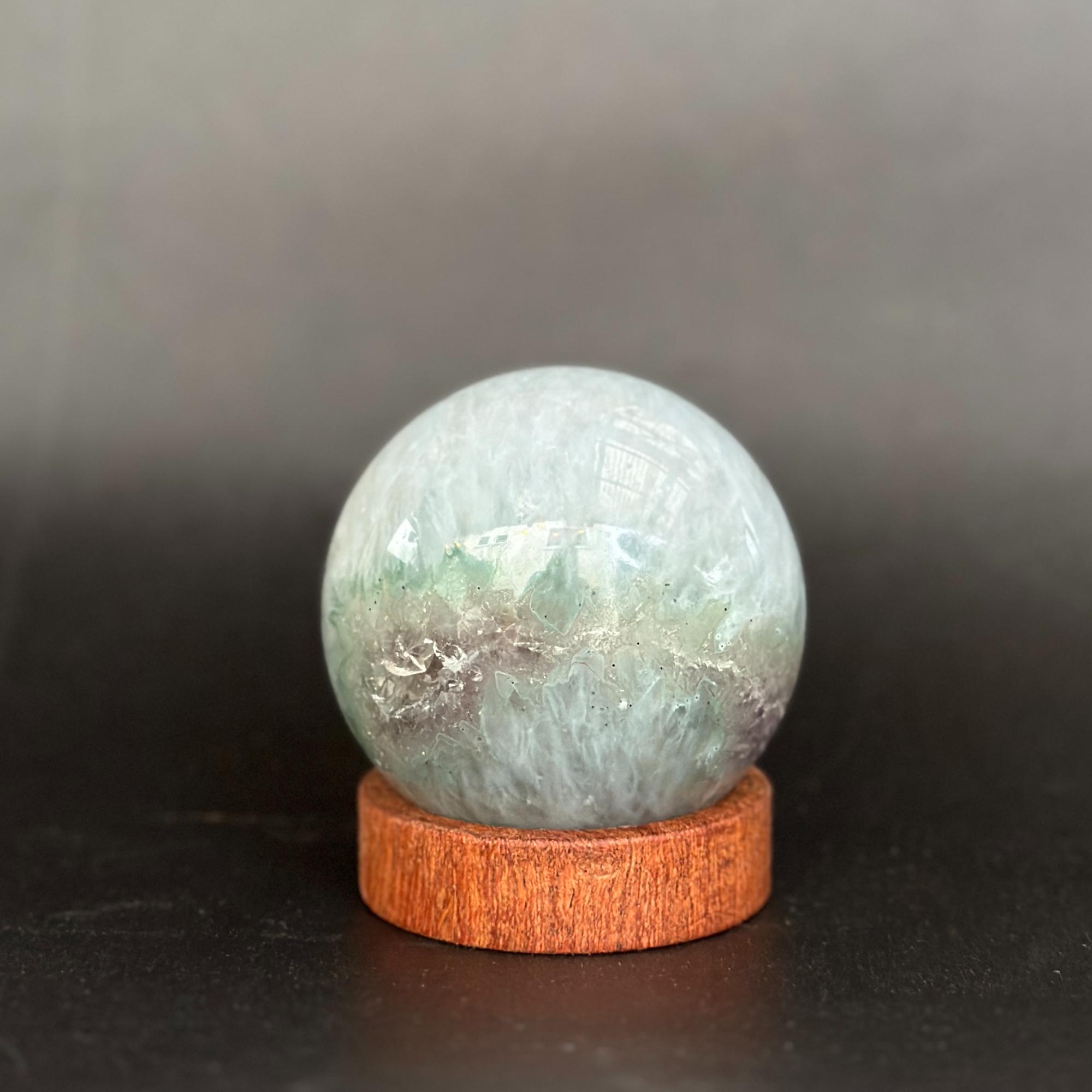 A polished round crystal ball with pale green and white marbled patterns sits on a small round wooden display stand against a dark gray background.