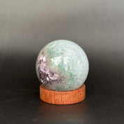 A polished crystal sphere with green, purple, and white marbling, displayed on a small round wooden base against a dark gray background.