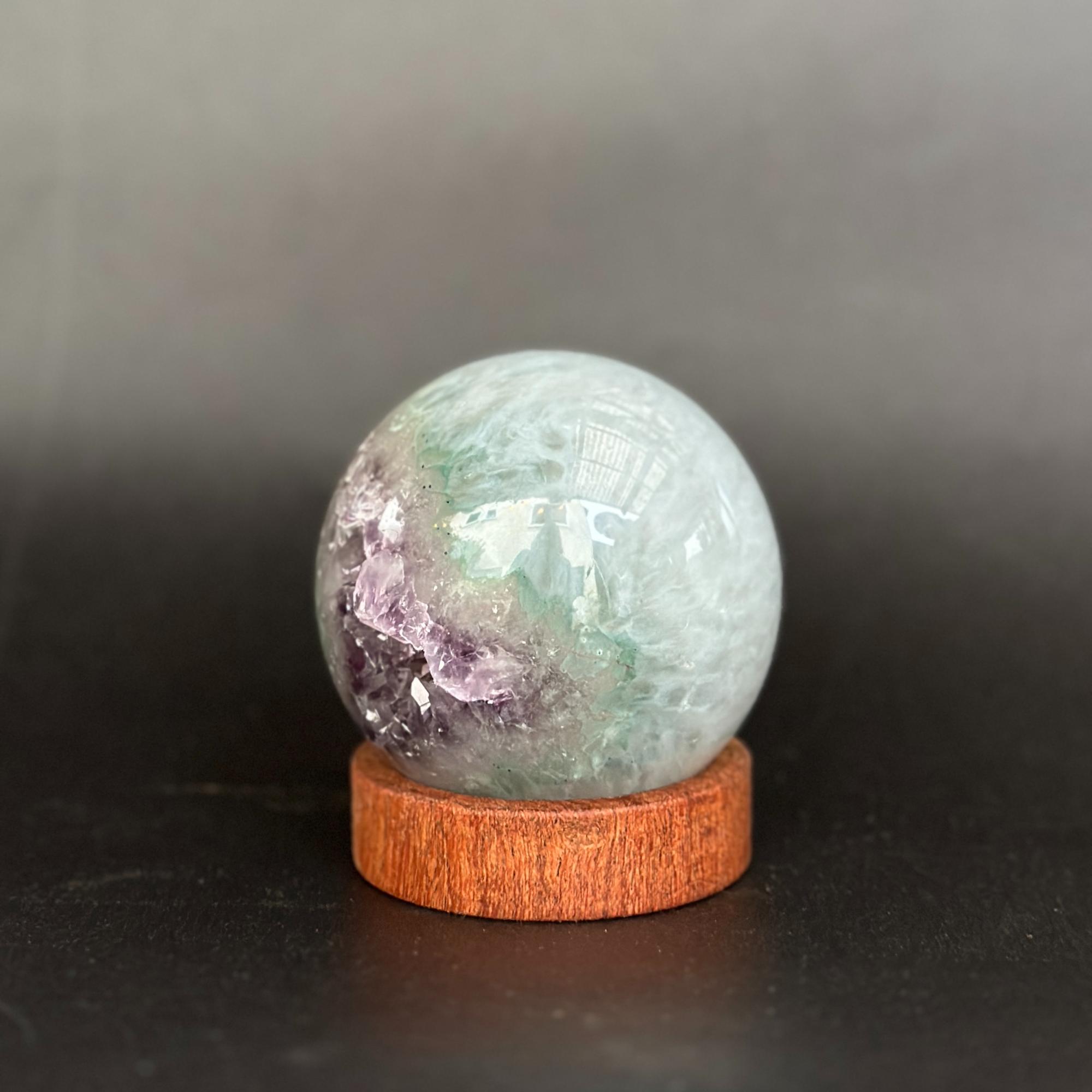 A polished crystal sphere with green, purple, and white marbling, displayed on a small round wooden base against a dark gray background.